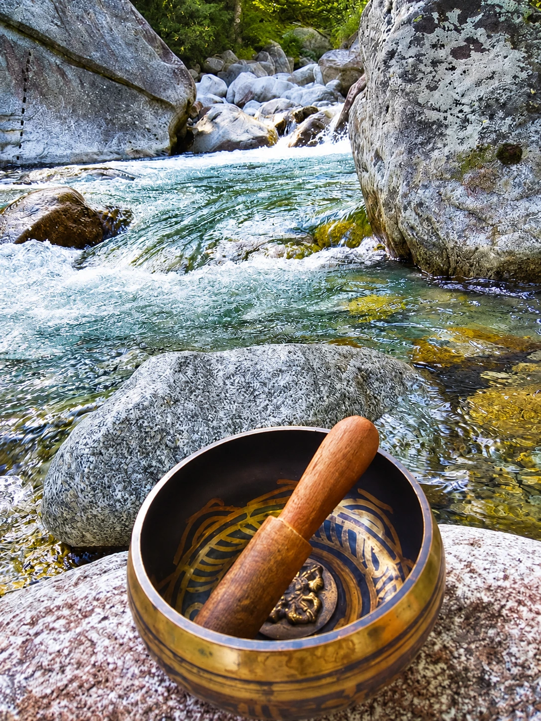 A singing bowl and wooden mallet resting on a rock beside a stream